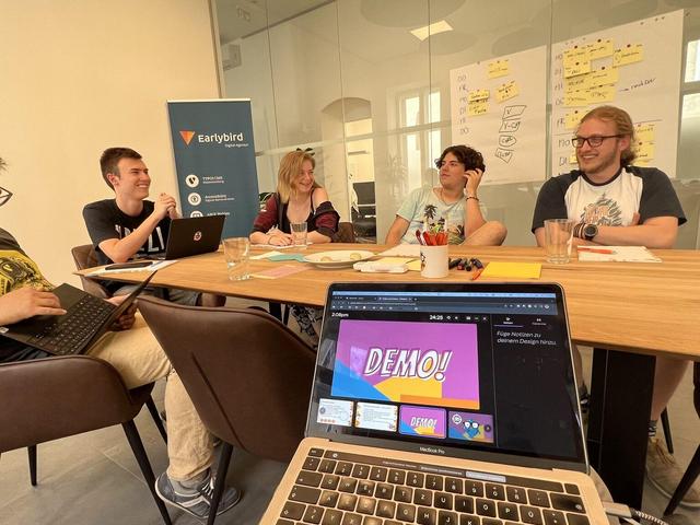 Blick über ein Laptop auf den Besprechungstisch. Leute arbeiten an einer Demo.