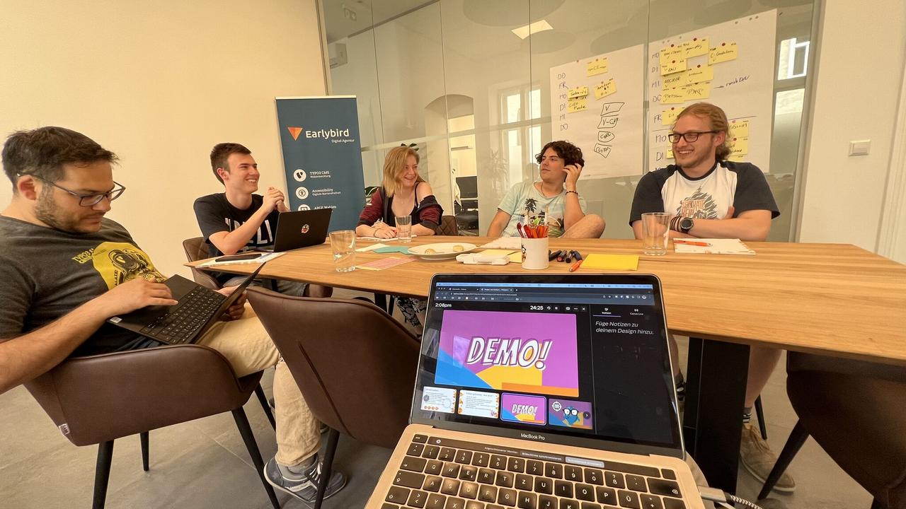 Blick über ein Laptop auf den Besprechungstisch. Leute arbeiten an einer Demo.