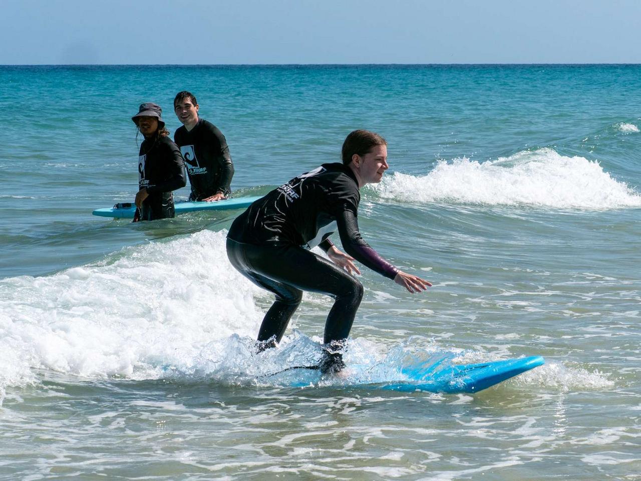 Nico auf dem Surfbrett am Wasser, dahinter Alex und der Trainer, die ihr zusehen