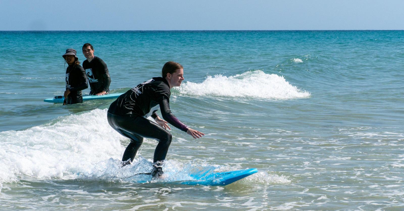 Nico auf dem Surfbrett, dahinter Alex und der Trainer im Wasser, die ihr beim Surfen zusehen